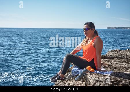 Donna latina, di mezza età, riposo, riconquista forza, mangiare, acqua potabile, dopo una sessione di ginnastica, indossando la parte superiore arancione, bruciando calorie, mantenendosi in forma, Foto Stock