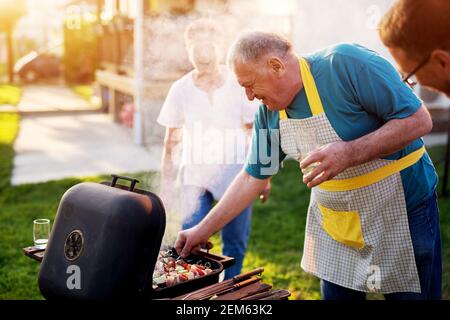 L'uomo anziano sta ispezionando attentamente se prende la carne fuori dalla griglia mentre sua moglie sta in piedi accanto a lui e ridendo. Foto Stock