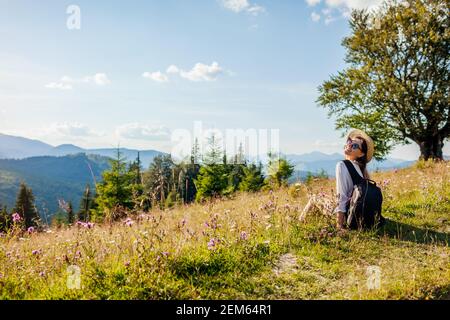 Viaggiare in Ucraina estate. Gita ai Monti Carpazi. Felice donna turista seduta in fiori dopo escursioni con zaino godendo la natura Foto Stock