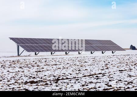 Pannelli solari montati a terra in campo agricolo coperto di neve in giornata nuvolosa. Concetto di energia rinnovabile, risparmio delle bollette di servizio e generazione di elettricità Foto Stock