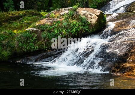 Corrente d'acqua che scorre intorno a formazioni rocciose ricoperte di muschio della cascata di Ipiranguinha formando una piscina naturale dorata proprio sotto. Foto Stock