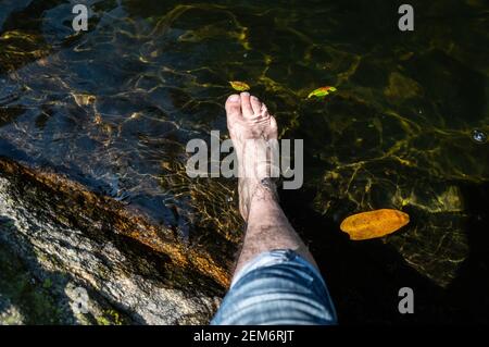 Gamba maschile indossando pantaloni jeans trainati blu e piede immerso in acque cristalline della piscina naturale dorata formata dalla cascata Ipiranguinha. Foto Stock