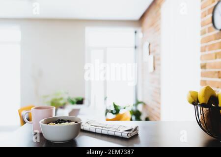 Tazza di caffè rosa, ciotola con kiwi e banana tritati di frutta tropicale, mirtilli, cucchiaio sul bancone bar in elegante cucina loft. Sfondo sfocato. Hig Foto Stock