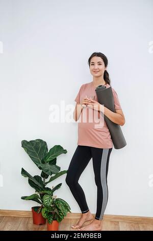 Donna incinta sorridente al club di yoga, tenendo il tappetino arrotolato, in posa per una foto Foto Stock