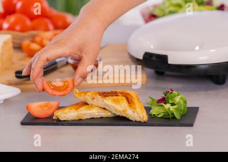 Mano femminile mettendo una fetta di pomodoro su un vassoio di ardesia con un formaggio alla griglia e un sandwich di prosciutto tagliato a metà. Messa a fuoco selettiva e primo piano. Concetto di cibo. Foto Stock