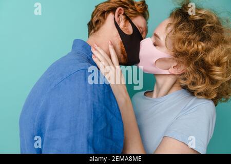 Primo piano di una giovane coppia attraente che indossa maschere di protezione baciare isolato su sfondo blu Foto Stock