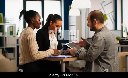 Entusiasta uomini d'affari diversi leggere il rapporto finanziario annuale seduta al tavolo in un moderno ufficio aziendale startup tenendo tablet e sorridendo. Team di imprenditori multietnici che lavorano in azienda Foto Stock