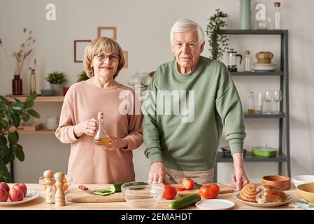 Ritratto di una coppia senior sorridente alla fotocamera mentre si è in piedi la cucina e la preparazione di insalata di verdure con olio d'oliva Foto Stock