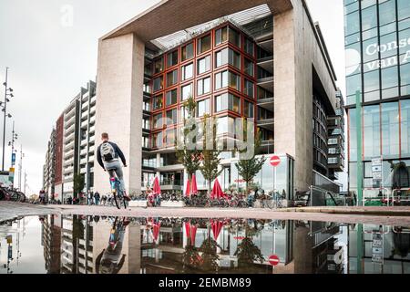 La scena stradale del centro di Amsterdam si riflette in un pozze d'acqua moderno edificio di appartamenti e turisti in bicicletta su strade deserte quartiere specchiato Foto Stock