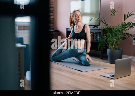 Giovane donna rossa flessibile che lavora, facendo esercizi di stretching su tappetino yoga tramite computer portatile Foto Stock