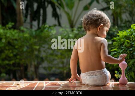 un affascinante bambino si siede con un giocattolo sui gradini di una casa sotto la luce del sole. vista posteriore. Foto Stock