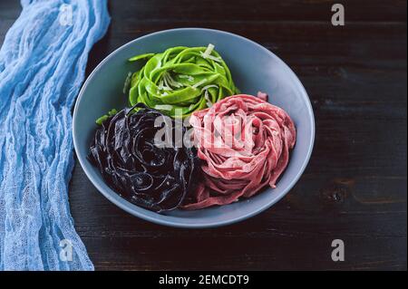 tre deliziose paste, pasta nera, pasta verde e fucsia. piatto grigio su fondo ligneo. Foto Stock