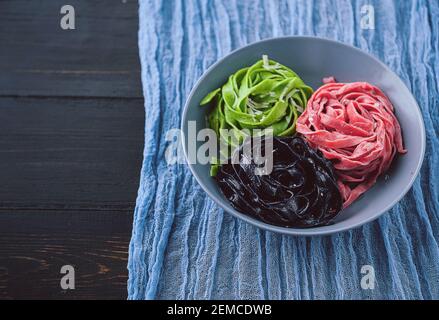 tre deliziose paste, pasta nera, pasta verde e fucsia. piatto grigio su fondo ligneo. Foto Stock