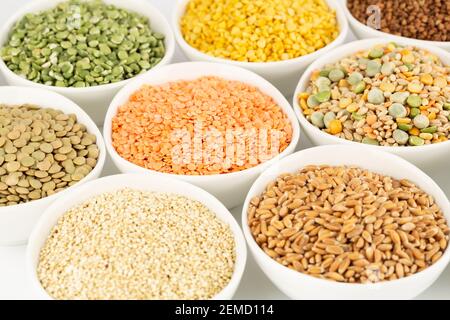 La raccolta di diversi tipi di groat nelle ciotole in ceramica bianca. Piselli spezzati, lenticchie colorate, grano saraceno, grani di grano saraceno, quinoa. Foto Stock
