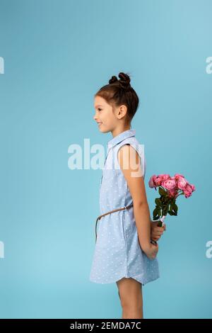 bambina piccola ragazza che tiene bouquet di rose dietro la schiena su sfondo blu. vista laterale Foto Stock