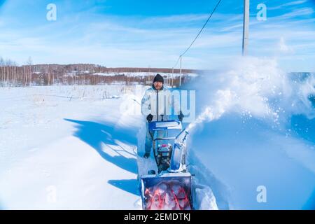 Un uomo in una giacca bianca rimuove la neve da a. strada rurale con un snowblower in inverno dopo una nevicata Foto Stock