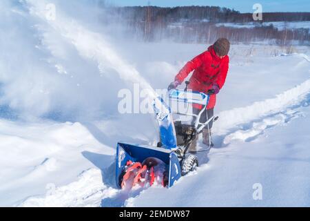 Una donna in una giacca rossa rimuove la neve da a. strada rurale con un soffiatore di neve blu in inverno dopo un nevicata Foto Stock