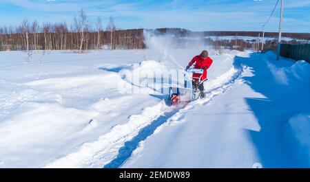 Una donna in una giacca rossa rimuove la neve da a. strada rurale con un soffiatore di neve blu in inverno dopo un nevicata Foto Stock