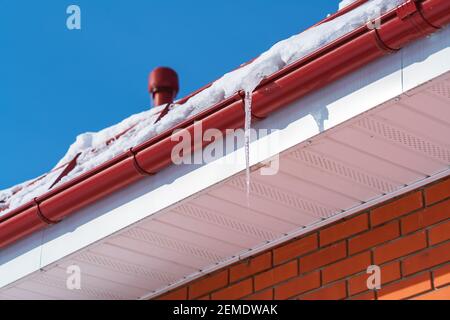 Un grande icicolo trasparente contro un cielo blu chiaro è sospeso da un tubo di scarico rosso in inverno alla vigilia di molla Foto Stock