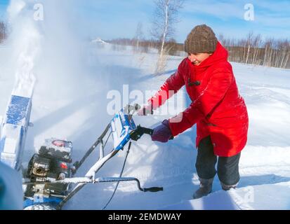 Una donna in una giacca rossa rimuove la neve da a. strada rurale con un soffiatore di neve blu in inverno dopo un nevicata Foto Stock