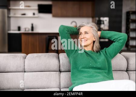 La donna anziana winsome riposa sul comodo divano a casa, affascinante donna anziana in abbigliamento casual messo le mani dietro la testa e sognare giorno in atmosfera rilassata, concetto di benessere Foto Stock