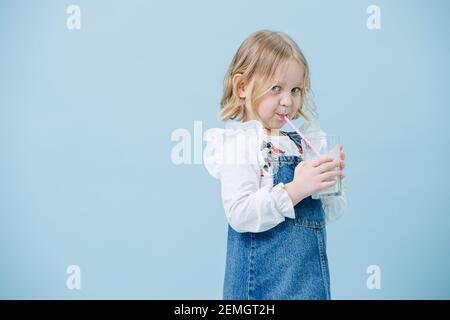 Perky bambina in tute jeans sorseggiando milkshake attraverso una paglia su blu Foto Stock