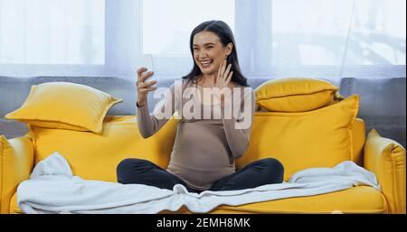 Donna incinta che agita la mano durante la chat video sullo smartphone a. casa Foto Stock