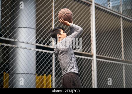 giovane basketball asiatico maschio che prende un colpo di salto sopra un campo recintato all'aperto Foto Stock