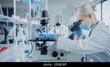 Laboratorio di Ricerca Medica moderna: Ritratto di scienziato femminile indossando maschera facciale guardando sotto microscopio, analizzando campioni di prova. Avanzate Foto Stock