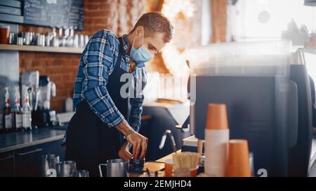 Il barista maschile nella maschera facciale sta premendo i fondi di caffè dopo la macinazione nel bar. Concetto di restrizioni di distanza sociale durante il Pandemic di Coronavirus Foto Stock