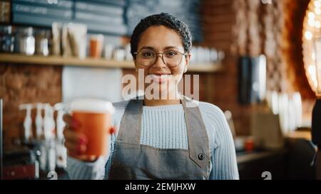 Bella barista femminile latino-americana con capelli corti e bicchieri è sorridente e tenere fuori una tazza di caffè take away in un caffè. Ritratto di felice Foto Stock