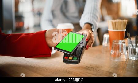 Primo piano di una mano femminile con cui tenere uno smartphone Modello schermo verde con tecnologia di pagamento NFC utilizzata per Pagando per il caffè da portare via in un Foto Stock