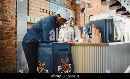 Beautiful Happy Latin Barista serve ordine a un corriere di consegna cibo raccolta sacchetto di carta con pasticcini da un ristorante Cafe. Delivery Guy mette Foto Stock