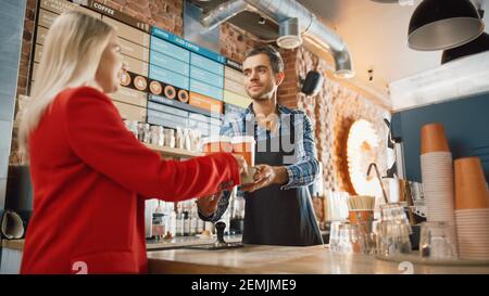 Shot in Modern Hipster Cafe: Bel barista sorridente in camicia a scacchi passa il portabicchieri di cartone con due tazze da caffè take away a una giovane femmina Foto Stock