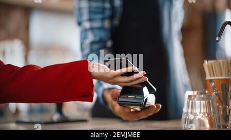 Primo piano di una mano femminile tenere uno smartphone con una tecnologia di pagamento NFC utilizzata per pagare il caffè da portare via in un caffè. Il cliente utilizza Mobile Foto Stock