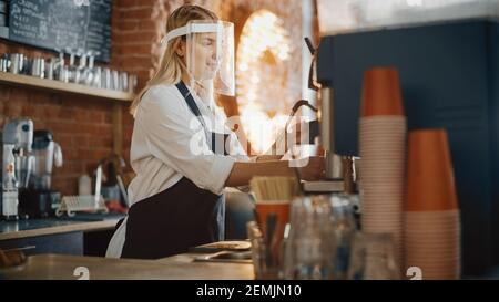 Bella giovane barista indossare Face Shield sta facendo una tazza di caffè fresco in un caffè. Bar dipendente che lavora nel Coffee Shop Restaurant. Sociale Foto Stock