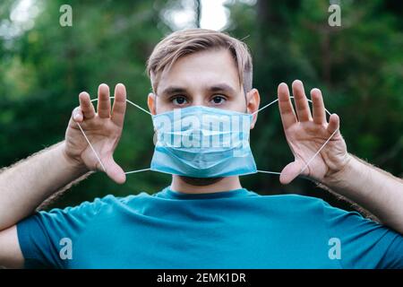 Uomo caucasico spaventato che tiene una maschera medica blu davanti al viso all'esterno. Maschera respiratoria monouso chirurgica come protezione contro il coronavirus Foto Stock