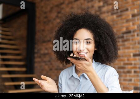 Giovane donna d'affari biraciale o un'imprenditore femminile con acconciatura afro che indossa abiti casual, seduto contro una parete di mattoni, tenendo il telefono cellulare, parlando, chattando e registrando un messaggio vocale Foto Stock