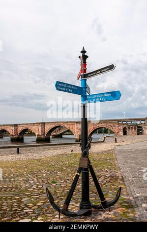 Un moderno cartello con due ancore attaccate alla sua parte inferiore Sezione si trova sui ciottoli di Berwick Quay con il vecchi e nuovi ponti dietro Foto Stock