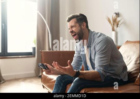 Vista laterale ritratto di giovane hipster sopportato ragazzo seduto sul divano nel soggiorno, tenendo il joystick, gridando a causa della partita perso video gioco, maschile addicted gamer sensazione di pazzo, gesturing Foto Stock