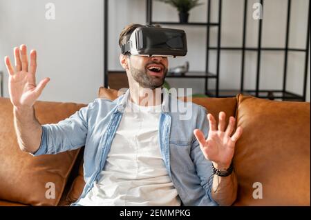 Un giovane e entusiasta ragazzo millenario con bocca aperta seduto sul divano e toccando l'aria con le mani, godendo di trascorrere il tempo libero in realtà virtuale a casa, giocando ai videogame con il moderno visore VR Foto Stock