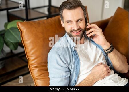 Primo piano di un giovane hipster sopportato sdraiato su un comodo divano ha risposto alla telefonata e ha avuto una piacevole conversazione, parlando con un amico tramite telefono cellulare, utilizzando l'applicazione di social media Foto Stock