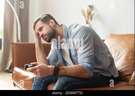 Vista laterale ritratto di giovane deluso hipster ragazzo, giocatore, o dipendente seduto sul divano nel soggiorno, tenendo il joystick, sensazione di stress e preoccupato a causa del fallimento del videogioco Foto Stock