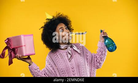Donna afroamericana in berretto di partito che tiene il regalo e lo champagne flacone isolato in giallo Foto Stock