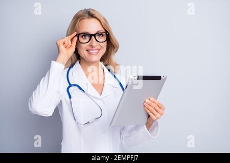Foto di lucido dolce donna medico indossare camice braccio bianco occhiali che tengono il dispositivo moderno spazio vuoto isolato sfondo grigio Foto Stock