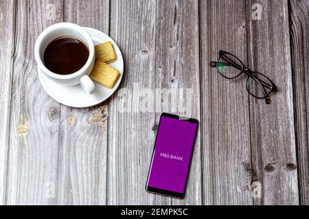 Un telefono cellulare o cellulare su un tavolo di legno Con l'app M&S Bank aperta accanto a un caffè e bicchieri Foto Stock