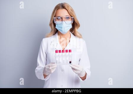 Foto di una donna chimica matura in occhiali e maschera facciale tenere le provette con il liquido all'interno isolato di colore grigio sfondo Foto Stock