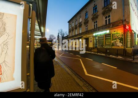 Cracovia, Polonia - 20 febbraio 2021: Retro di una donna in panni invernali e cappellino in piedi in una stazione di autobus in attesa di un arrivo in autobus contro polacco commerci Foto Stock