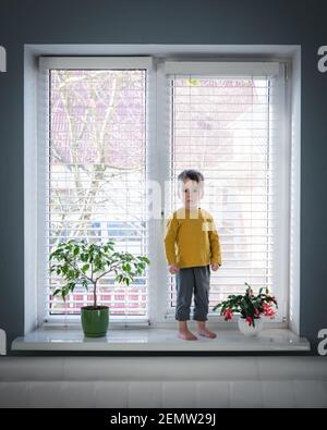 Piccolo ragazzo di solitudine stare sul davanzale vicino alla grande finestra nella sua stanza. Concetto di infanzia Foto Stock