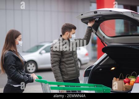 Giovane coppia in maschere di carico borse in tronco dopo supermercato shopping durante la quarantena. Uomo con sua moglie nel parcheggio all'aperto Foto Stock
